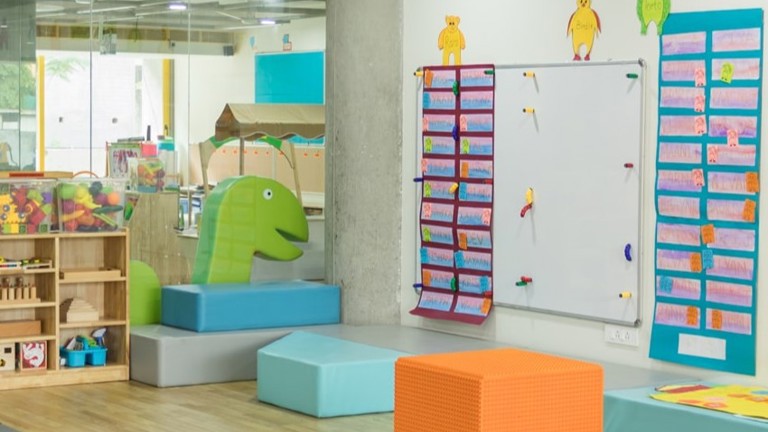 A vibrant daycare classroom with colorful toys, including a green dinosaur-shaped foam block, shelves of books and toys, and a whiteboard decorated with children's work.