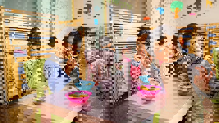 Group of young children eating snacks at a pink table, with one child in the foreground wearing an apron and looking playfully at the camera. Bunk beds are visible in the background.