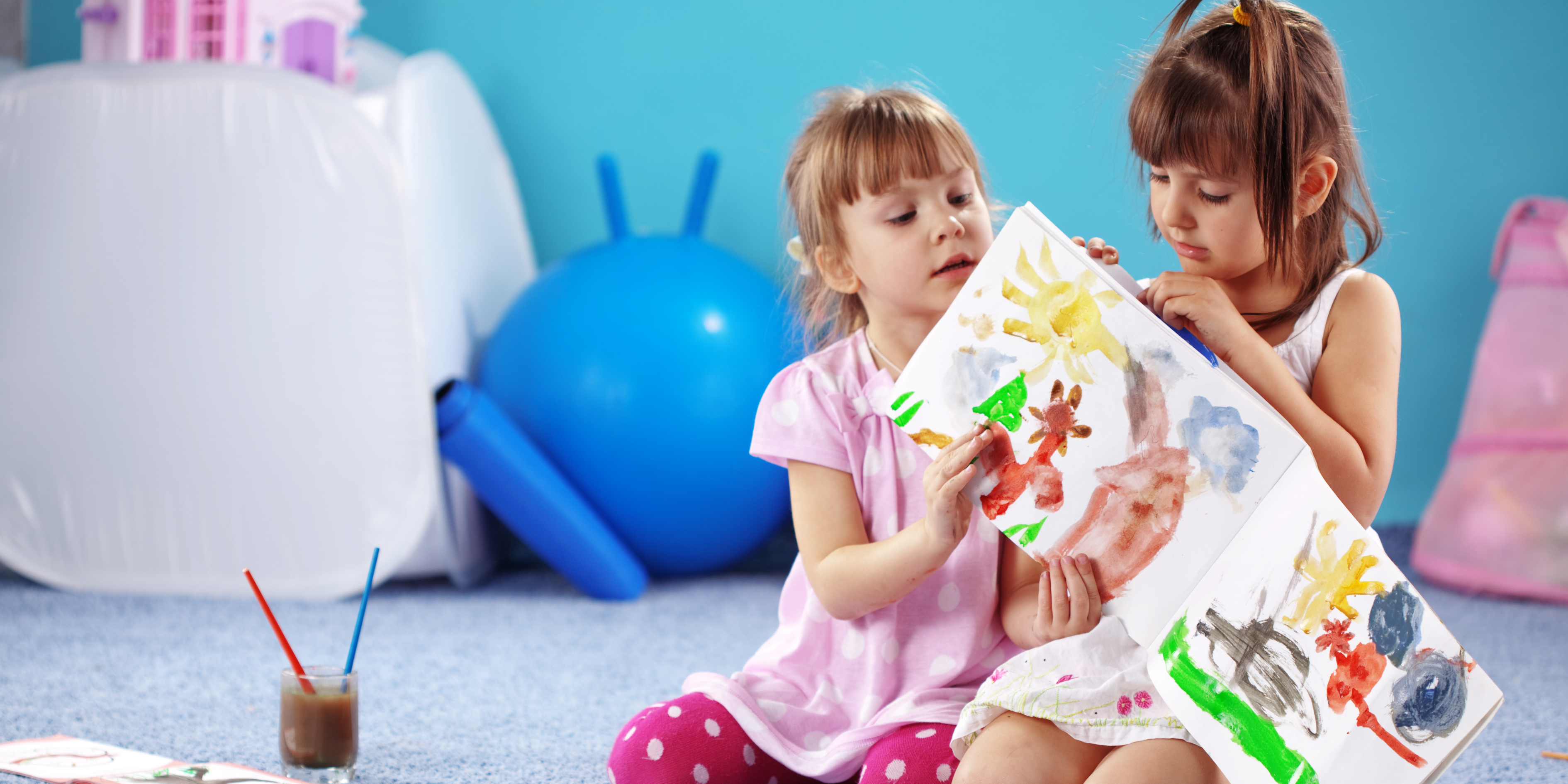 Two girls with brown hair, in pink and white dresses, sit on a gray carpet looking at a painting. A blue wall and exercise ball are in the background.