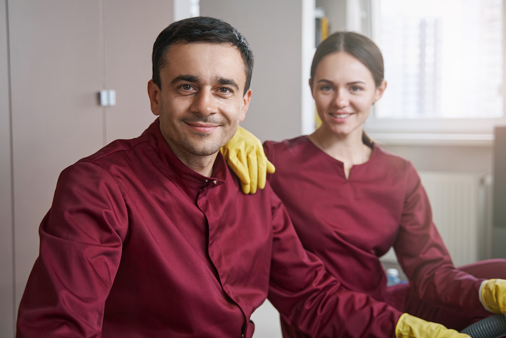Smiling man and woman in matching dark red shirts and yellow gloves pose together, appearing to be professional cleaners or maintenance staff.