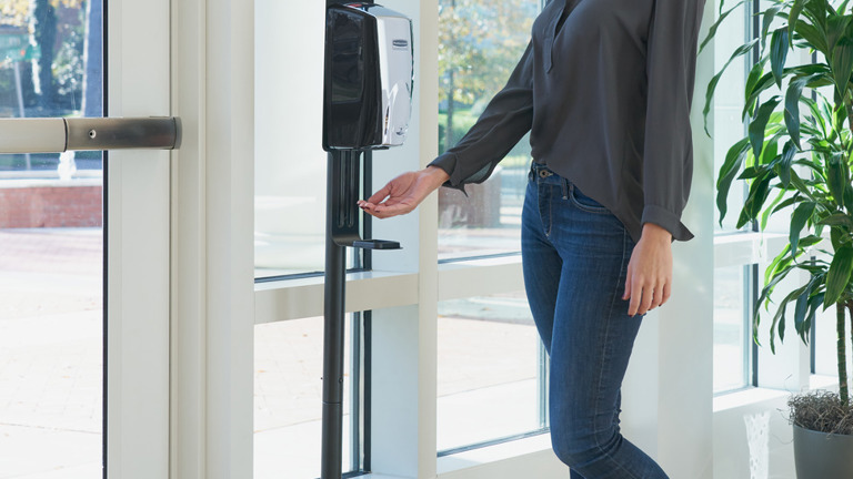 Person using a Rubbermaid Commercial AutoFoam Touch-Free Dispenser on a floor stand near a glass doorway