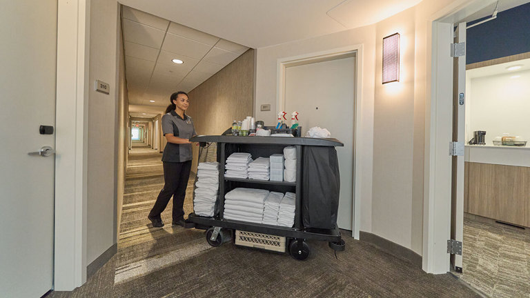 Female housekeeper pushing a Rubbermaid Executive Tradifilled cart with fresh towels and supplies down a hotel hallway.