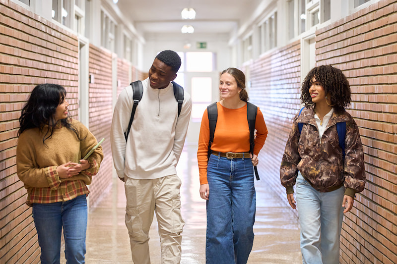 Four young adult students walking side by side in a school hallway, chatting and smiling with backpacks on.