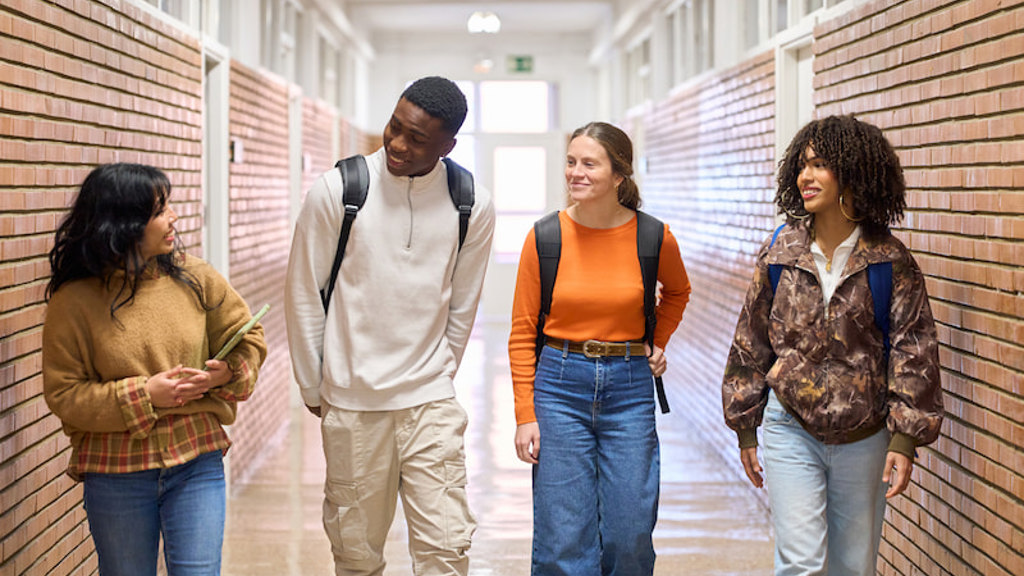 Four young adult students walking side by side in a school hallway, chatting and smiling with backpacks on.