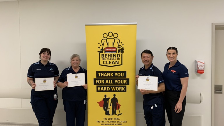 Four employees in dark blue uniforms holding certificates, standing in front of a yellow Rubbermaid Commercial Products banner thanking staff for their hard work.