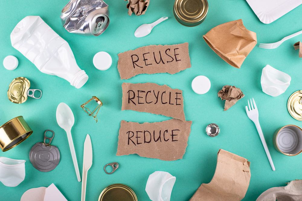 Top-down view of recyclable and non-recyclable waste—plastic bottles, aluminum and tin cans, paper, and utensils—surrounding torn cardboard labeled 'REUSE,' 'RECYCLE,' and 'REDUCE.'
