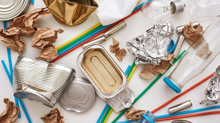 Assorted recyclable waste including crumpled aluminum and tin cans, glass and plastic bottles, a light bulb, and plastic straws on a white surface.
