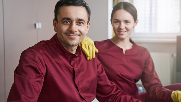Portrait of a man and woman in work uniforms and yellow cleaning gloves, smiling at the camera in an office or commercial setting.