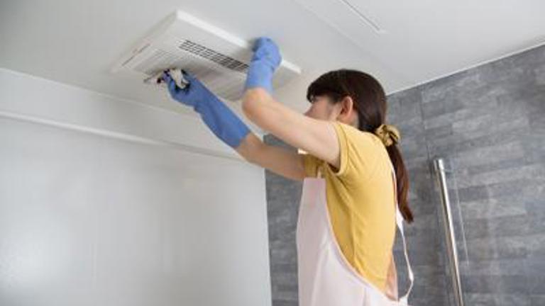 A woman wearing gloves and an apron is deep cleaning a ceiling fan